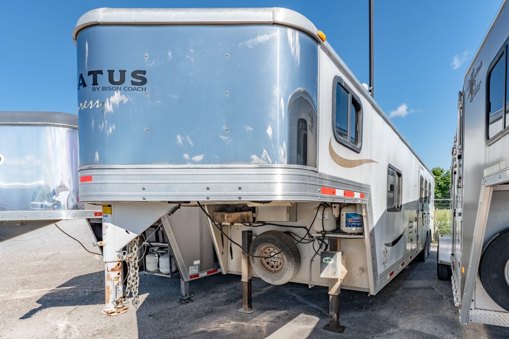 Silver-blue travel trailer parked on a lot, showing its front hitch, landing gear, and exposed storage area beneath the frame.