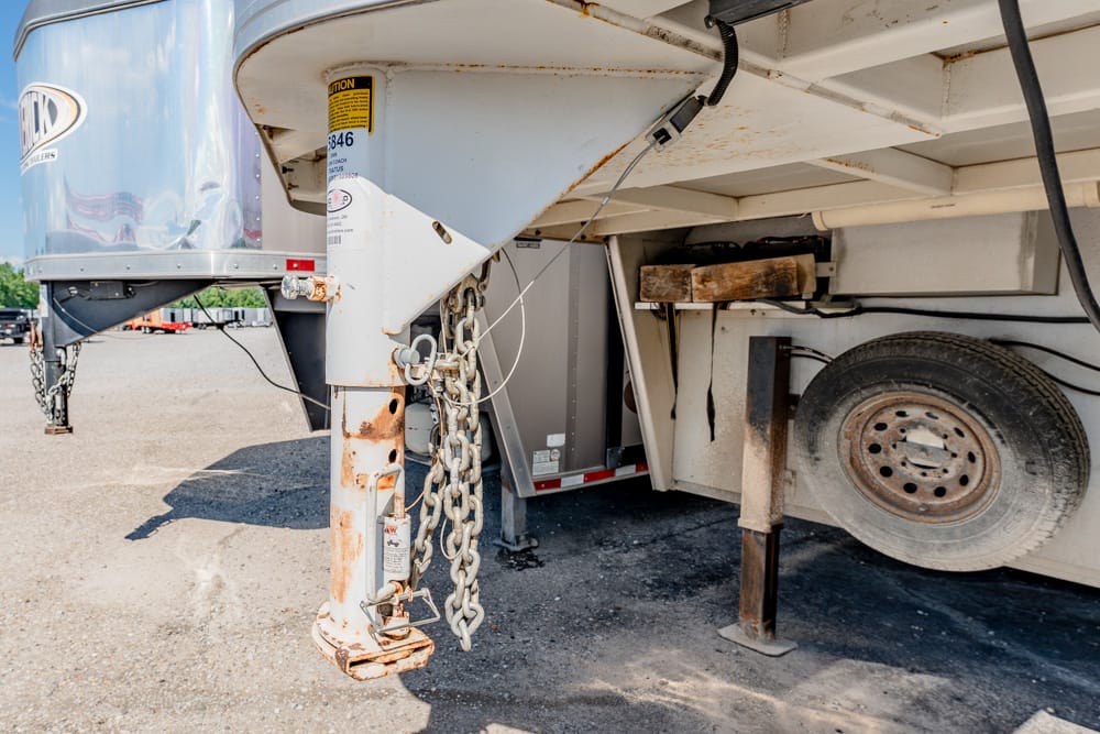 Close-up of a rusty trailer landing gear, chains, and support jack beneath a semi-trailer on a gravel lot, with a spare wheel nearby.