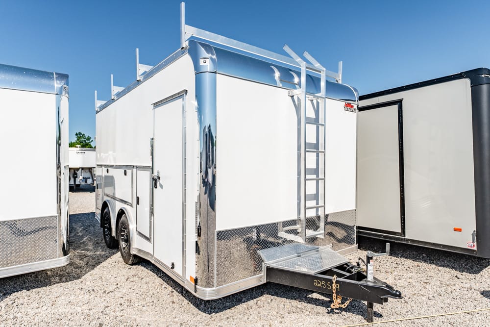 White enclosed cargo trailer with chrome trim and a side ladder, parked on a gravel lot beside other trailers.