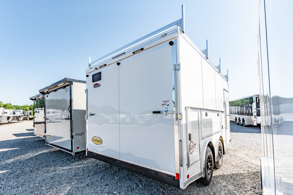 White enclosed utility trailer with dual rear doors parked on a gravel lot beside other trailers on a sunny day.