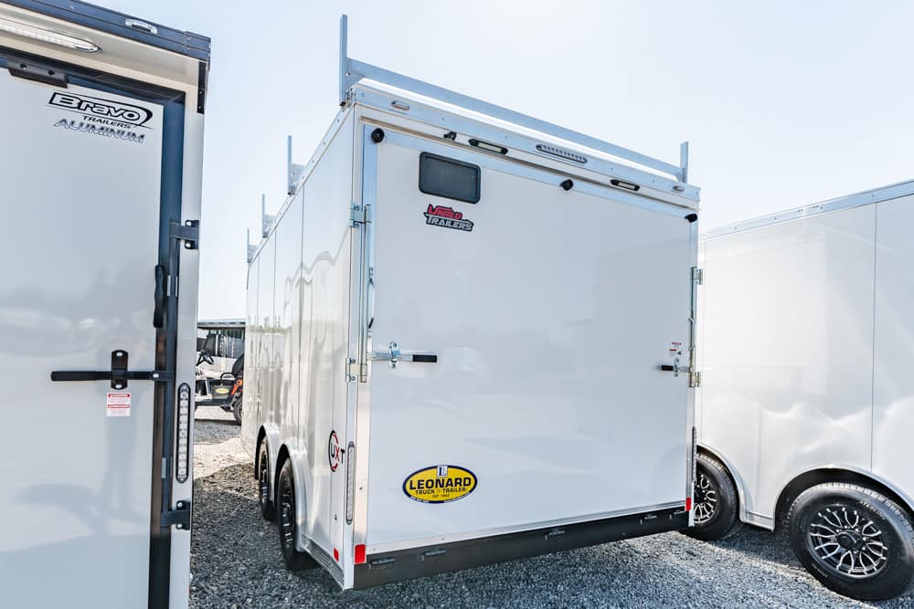 Rear view of a white enclosed utility trailer with double doors, parked on a gravel lot between two other trailers; small window on the back and various decals visible.