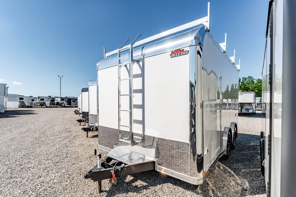 Silver enclosed trailer with a rear ladder and diamond-plate trim parked on a gravel lot, row of similar trailers in the background.