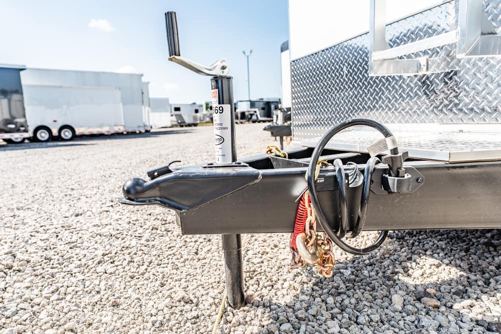 Close-up of a trailer hitch with a jack, safety chains, and lock cables on a gravel lot, ready for connection or securing a trailer adjacently parked equipment.