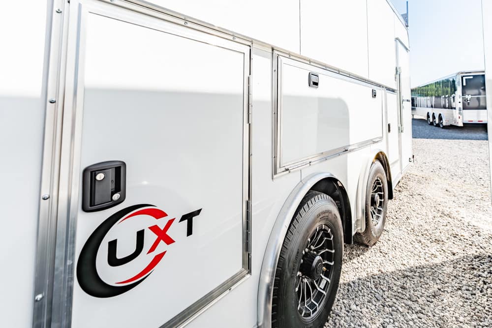 Side view of a white utility trailer with the UXT logo, parked on a gravel lot with visible storage doors and a rear wheel.**