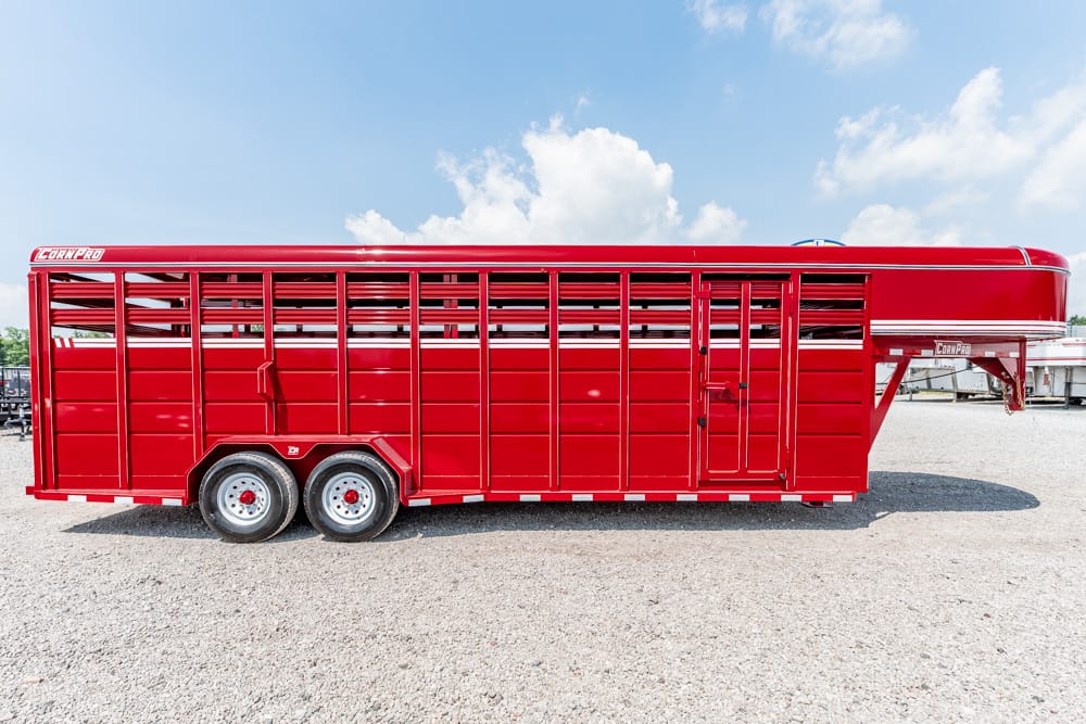 Red gooseneck livestock trailer with vented sides and twin axles on a gravel lot under a blue sky.