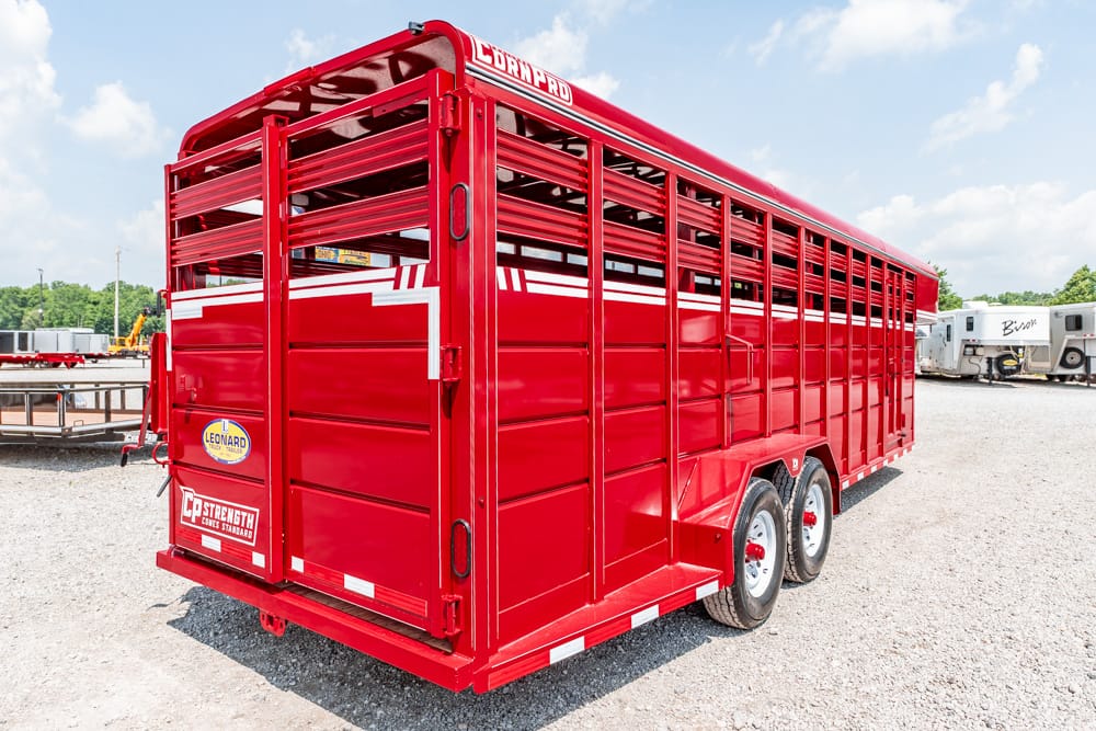 Red livestock trailer with multiple vents and a white stripe, parked on a gravel lot with other trailers in the background, two rear doors closed and dual axles visible.