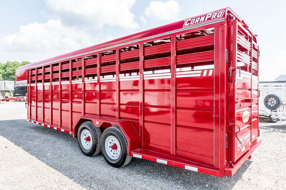 Red livestock trailer with vented side panels and dual rear wheels on a gravel lot at a farm show.