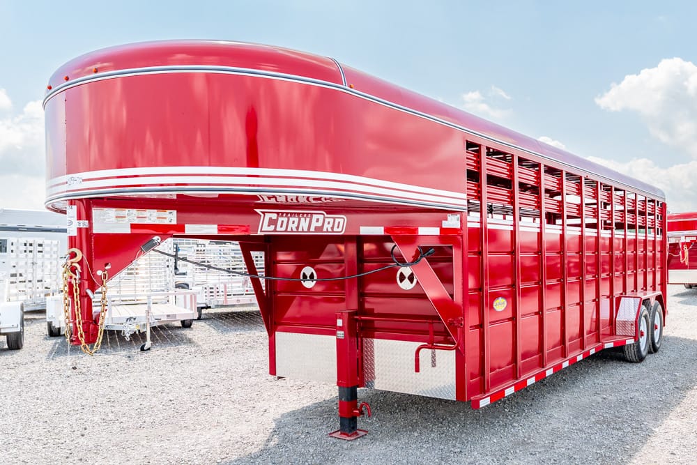 Red CornPro livestock trailer with vented side panels, parked on a gravel lot under a blue sky.