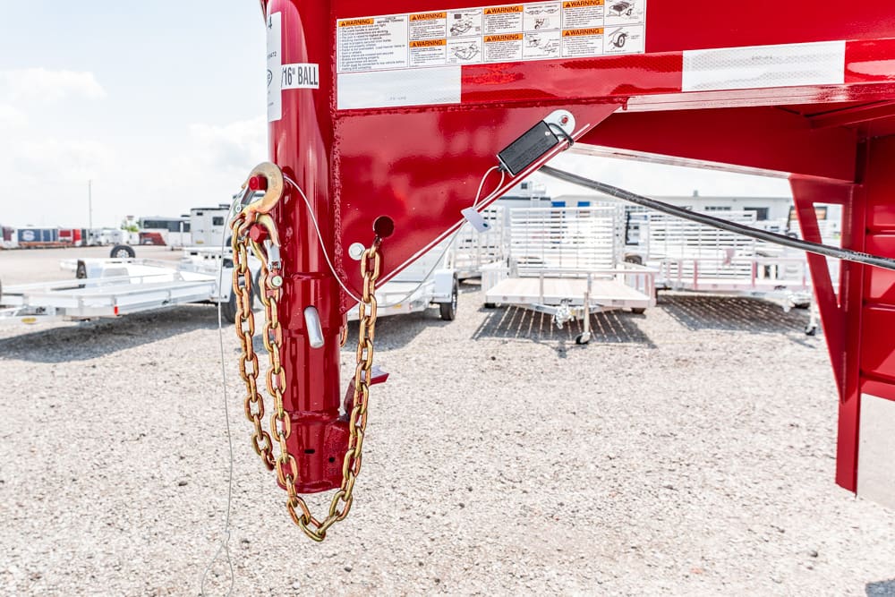 Close-up of a red trailer hitch with heavy gold chains and a padlock, in a gravel lot with other trailers in the background.