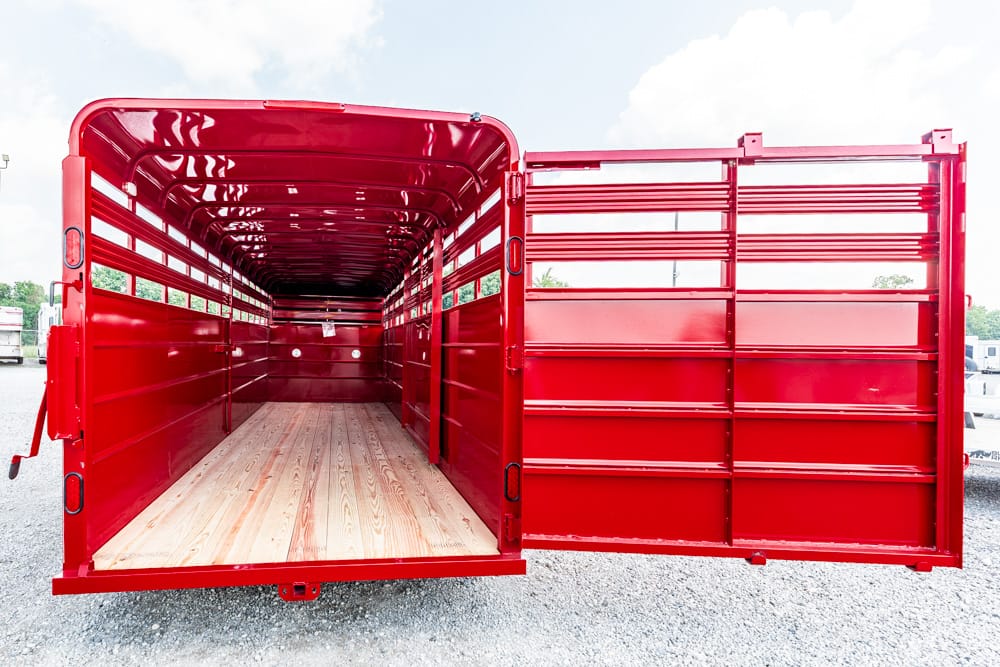 Open red trailer with wooden floor and metal side slats, ready for loading on a gravel lot.