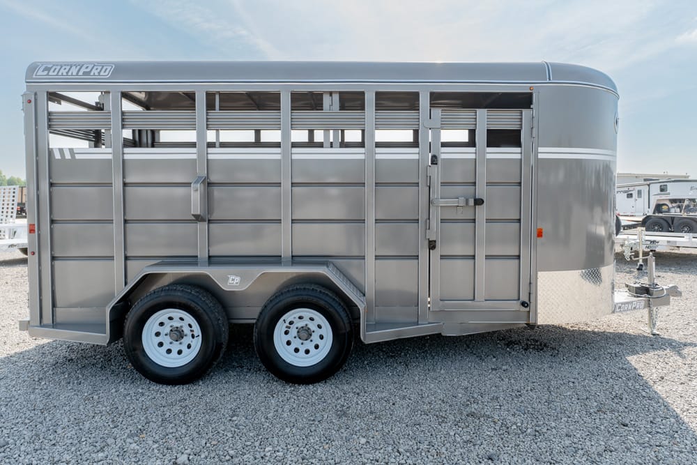 Silver two-axle livestock trailer with side access door and vented panels on a gravel lot, brand name visible on front corner edge.