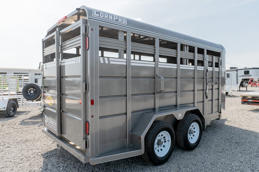 Gray CornPro livestock trailer with dual axles and a spare tire mounted on the rear, parked on gravel.