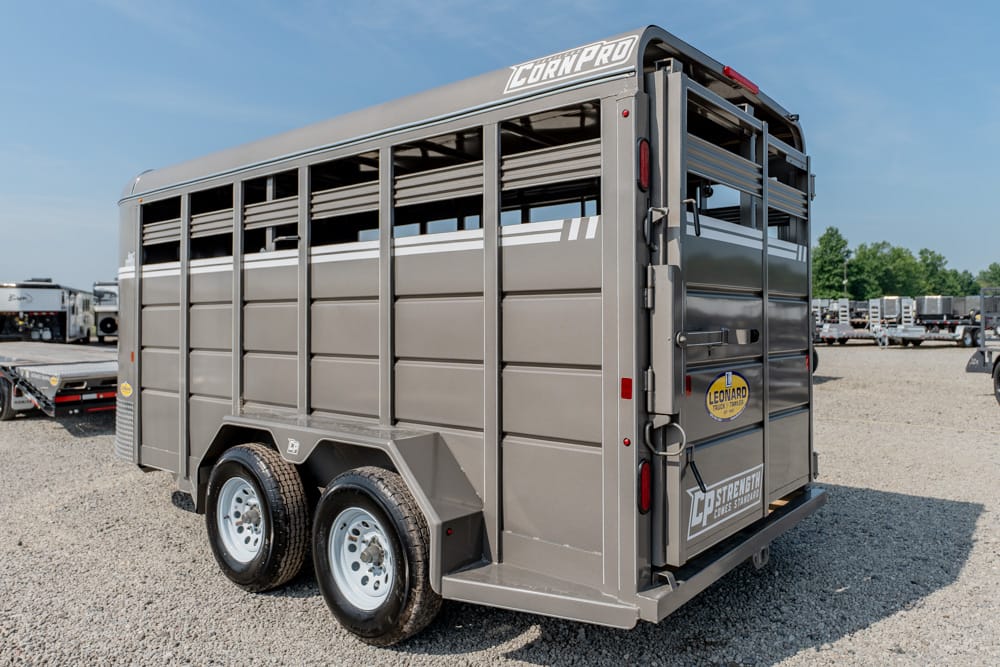 Gray metal livestock trailer with tandem wheels parked on a gravel lot, rear doors closed and brand decals visible.