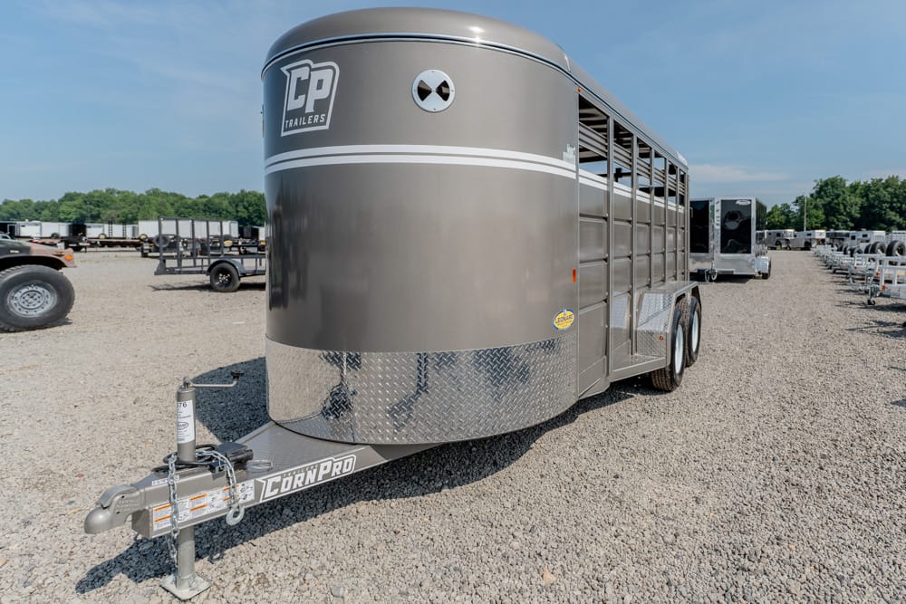 Gray aluminum stock trailer with dual axles and a rounded front sits on a gravel lot, CP Trailers logo near the top and diamond-plate rock guard at the front, connected to a CornPro hitch.
