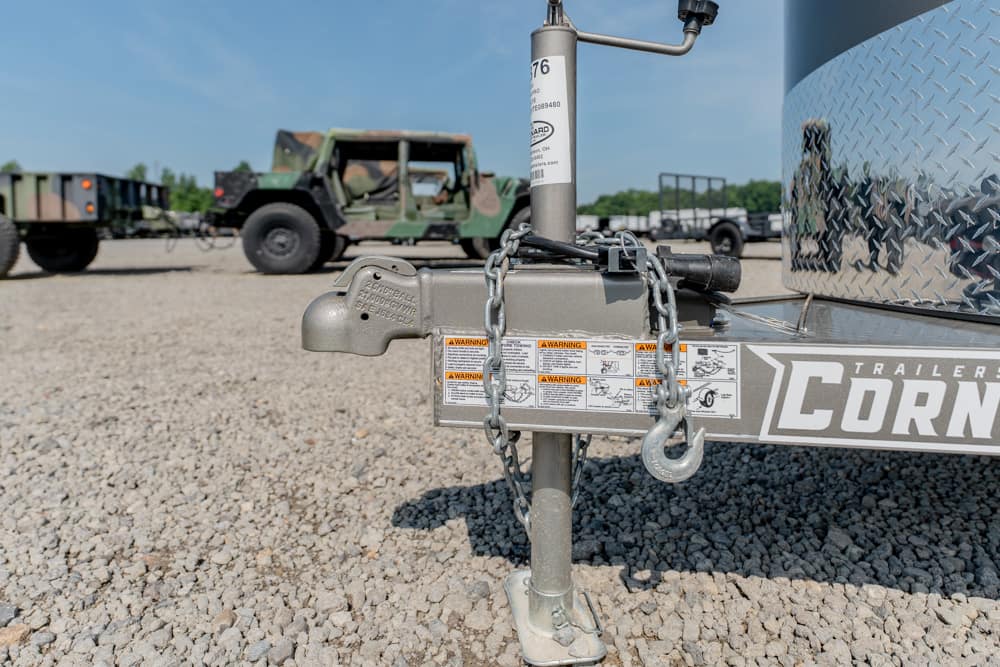 Close-up of a trailer tongue with a coupler, safety chains, and jack on gravel; military trucks in the background.