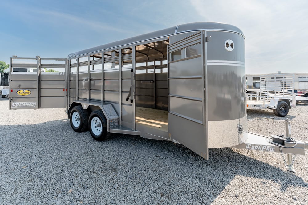 Two‑axle silver livestock trailer with open side doors, parked on a gravel lot.