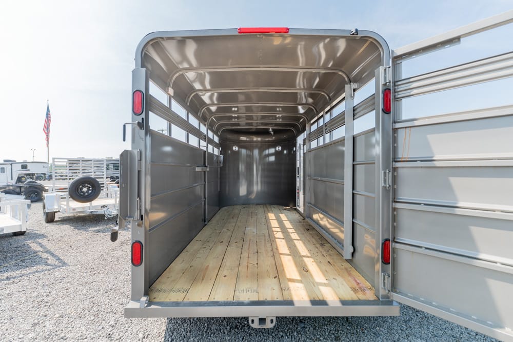Empty metal horse trailer interior with a wooden floor and side rails, rear doors open on a gravel lot.