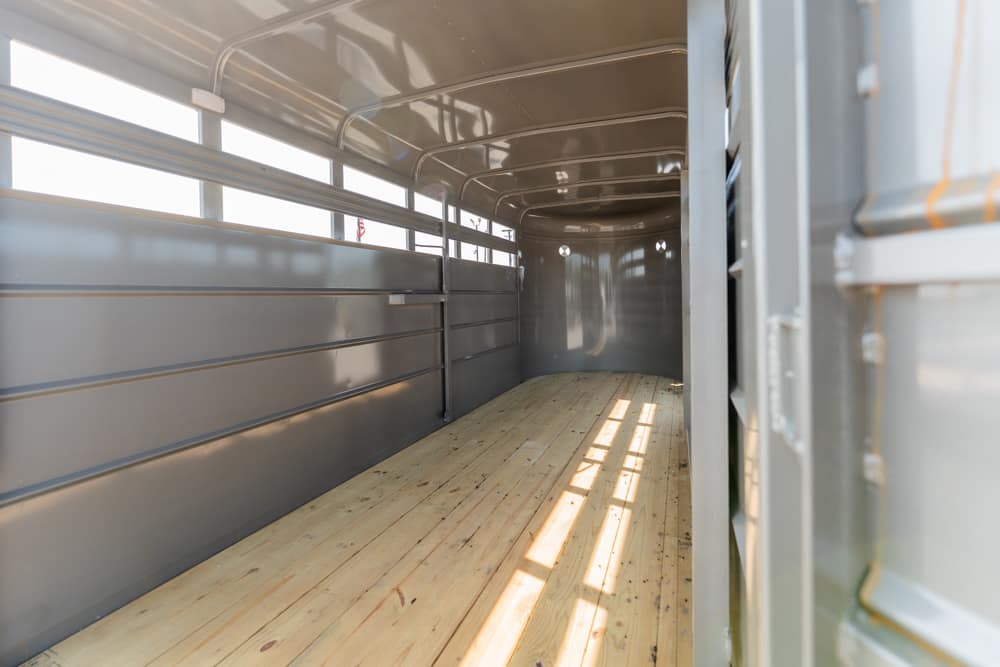 Interior of an empty metal livestock trailer with wooden plank floor and side rails, sunlight streaming through the windows.