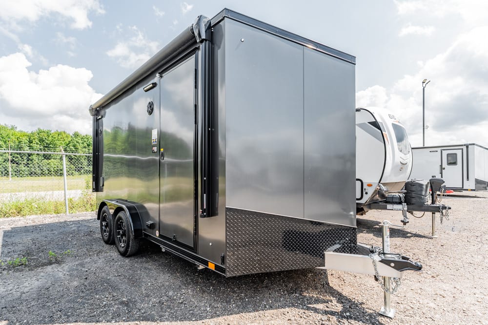 Enclosed dual-axle cargo trailer on a gravel lot, with a steel diamond-plate front and a hitch in the foreground; other trailers visible in the background.