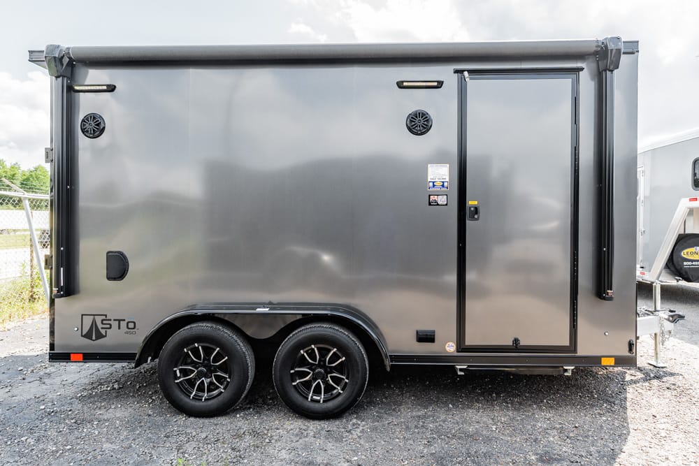 Gray enclosed cargo trailer with tandem wheels parked on gravel, side door closed and vents visible on the side