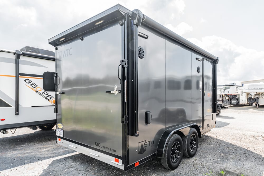Black enclosed cargo trailer with dual axles parked in a lot, other RVs visible in the background, brand logos on the sides.