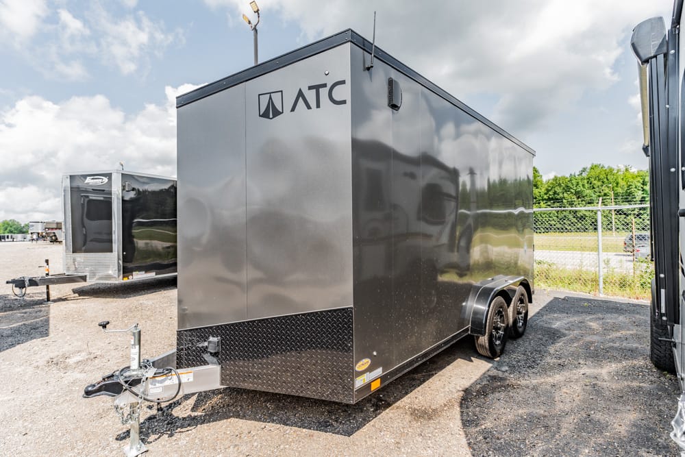 Black ATC enclosed cargo trailer with dual axles in a gravel lot, hitch at front, logo on side, reflective panels and diamond-plate rock guard at base.
