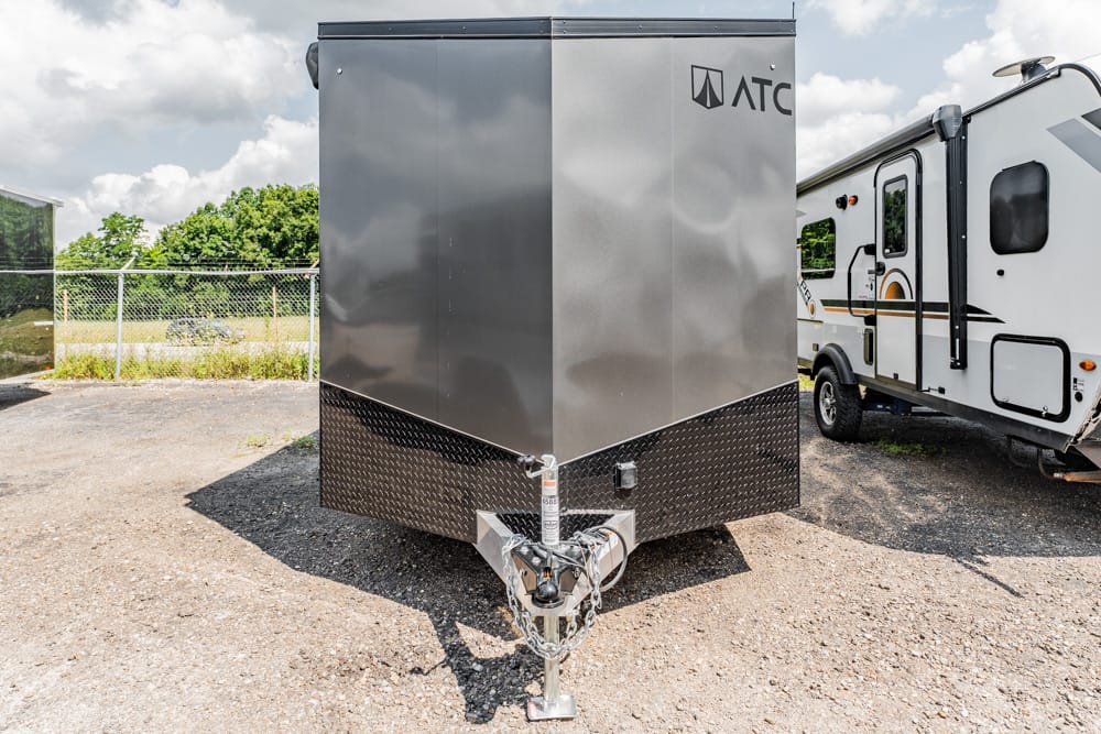 Front view of a gray enclosed cargo trailer with a diamond-plate lower front, ATC logo, and a trailer hitch with chains on a gravel lot near an RV.