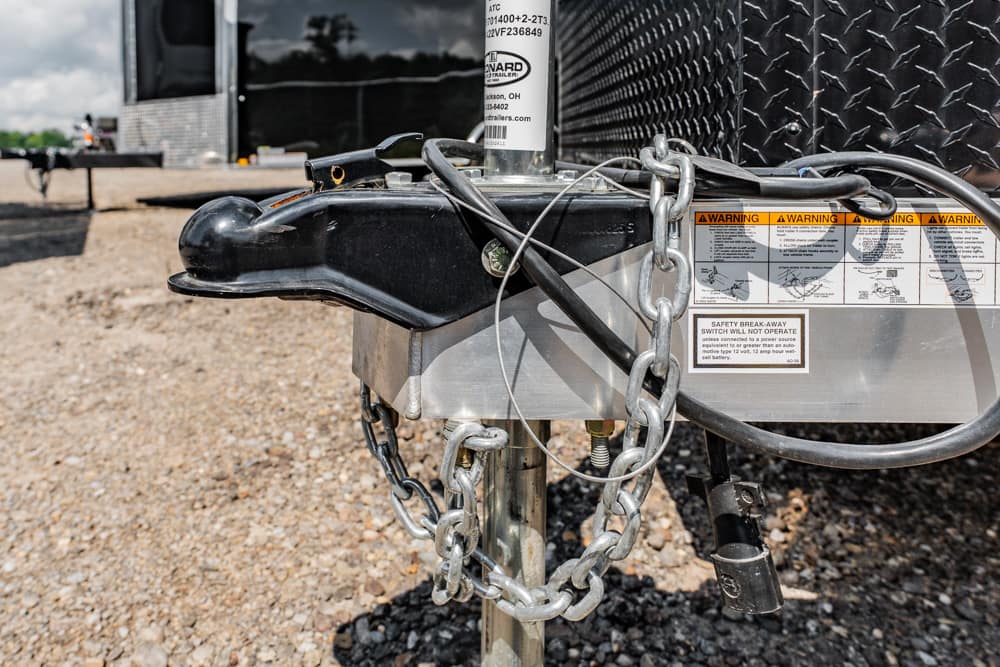 Close-up of a trailer tongue with safety chains and a padlock securing the hitch, warning labels on the side, on a gravel lot.