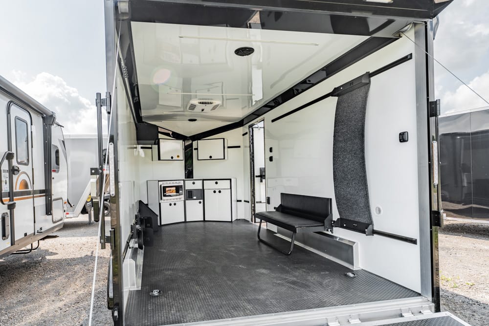 Interior of a converted utility trailer showing a compact kitchenette with white cabinets, a microwave, and a small fridge, plus a black bench and white walls with a curved gray wall panel on the right.