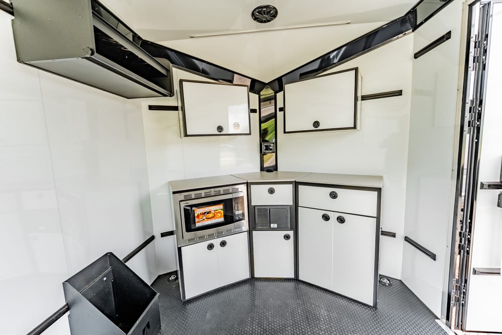 Compact kitchenette in a white trailer: microwave, small fridge, drawers, and upper cabinets.