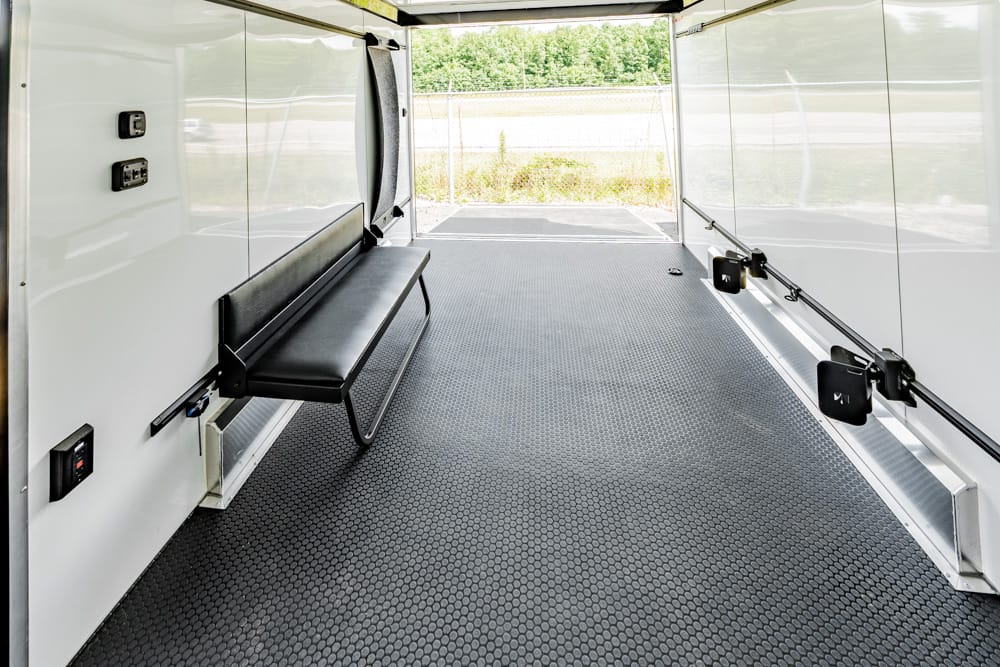 White cargo trailer interior with a black fold-down bench along the left wall and textured rubber flooring, open rear doors showing a fenced area outside.
