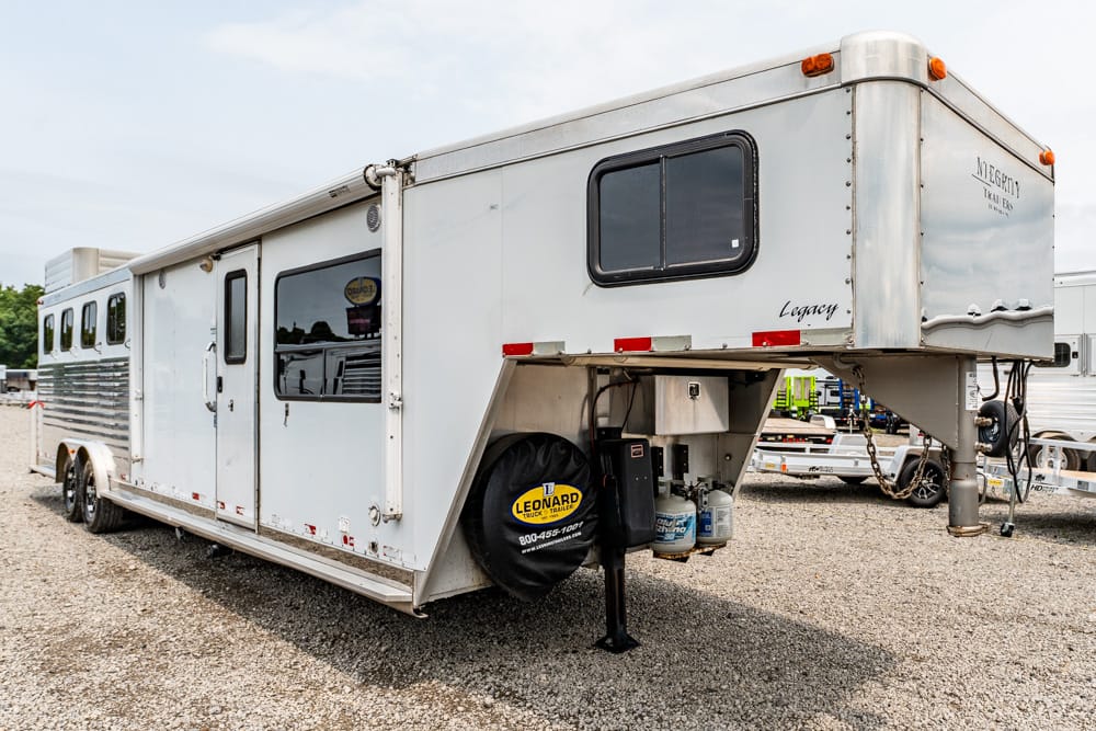 White gooseneck trailer parked on a gravel lot, with a spare tire, propane tanks, and dual axles visible