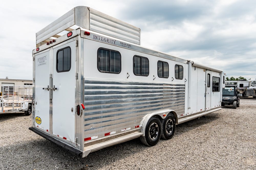 Silver horse trailer with ribbed aluminum siding, two axles, and small windows, parked on a gravel lot in a trailer yard.