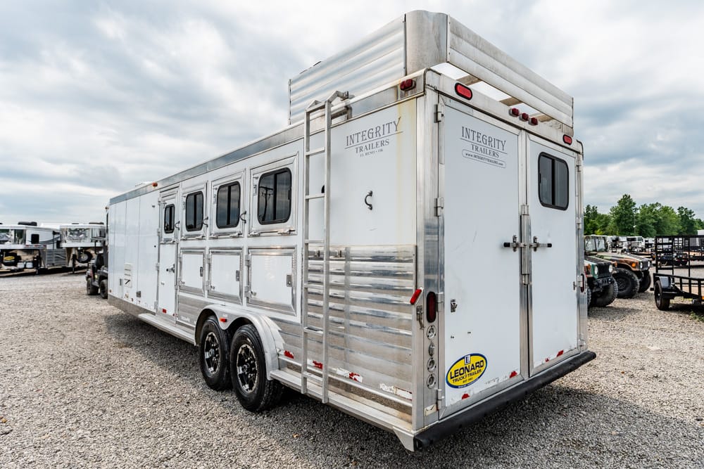 Silver aluminum livestock trailer with three small windows on the side and a rear double door, branded 'Integrity Trailers' in a gravel lot.