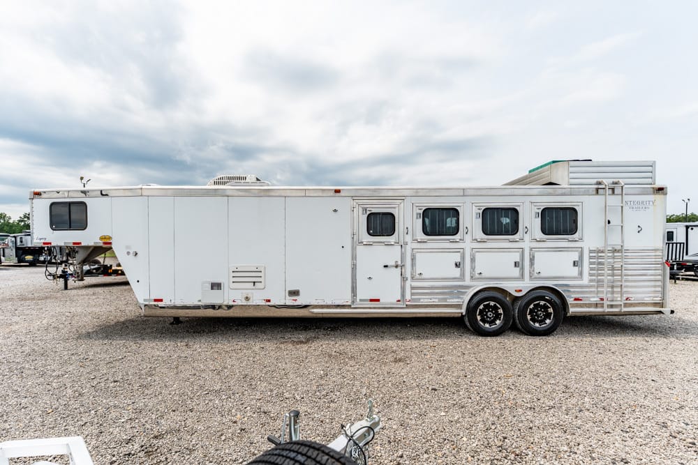 White horse trailer with several windows and storage compartments, parked on a gravel lot.
