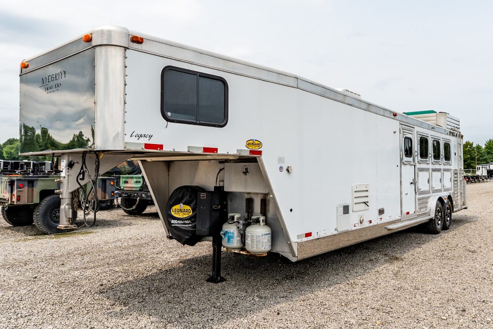 White long fifth-wheel RV/travel trailer with a front bedroom window and propane tanks at the hitch, parked on a gravel lot in a trailer yard.