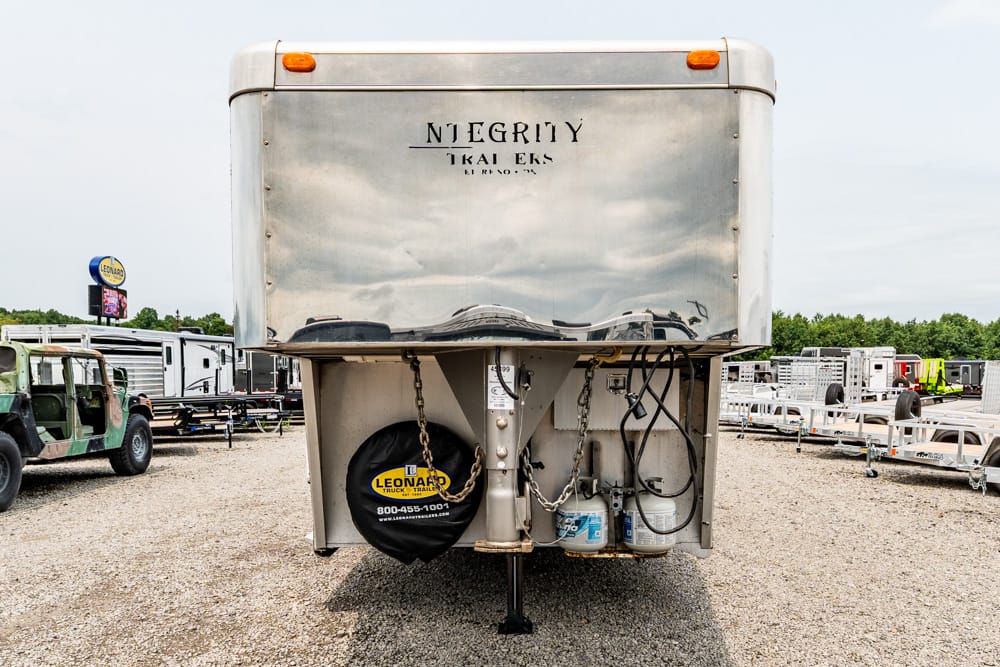 Front of a large aluminum trailer with a spare tire and propane tanks under the hitch in a gravel lot.