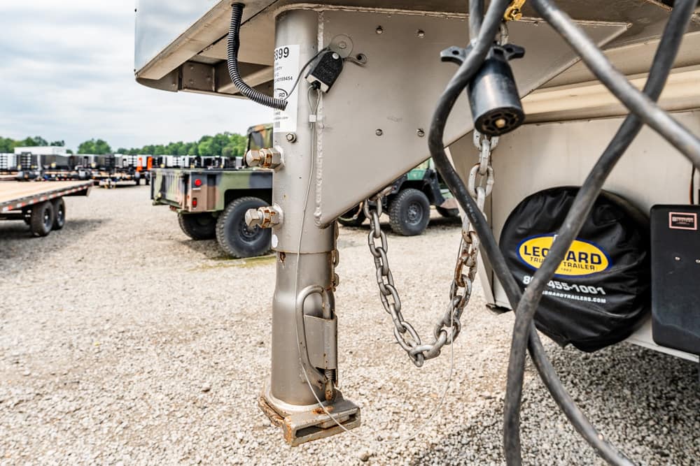 Close-up of a trailer hitch with safety chains and jack stand in a gravel lot with other trailers in the background.