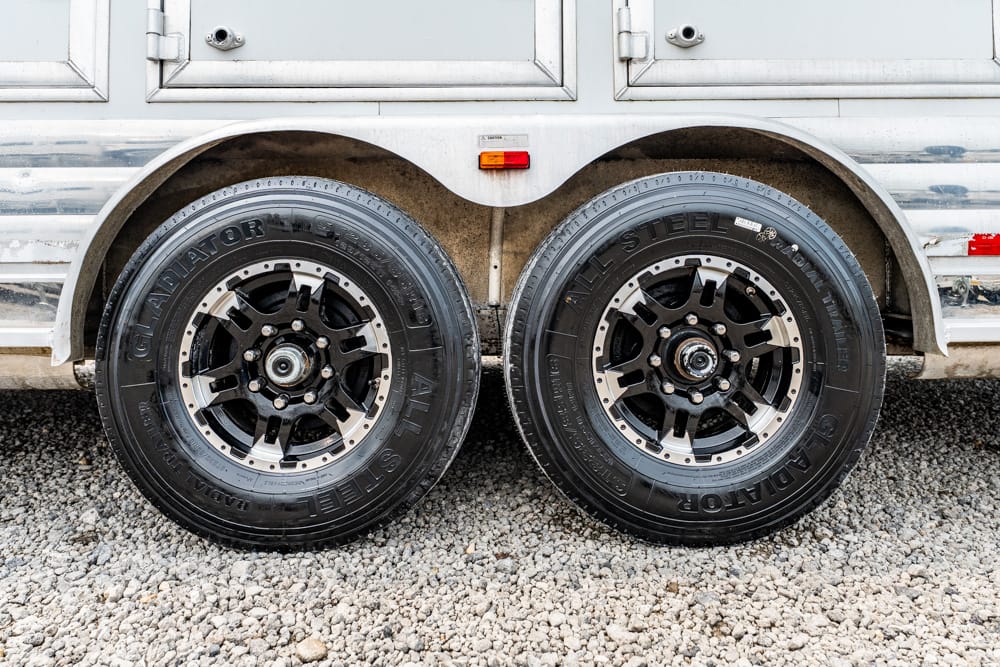 Two black alloy trailer wheels with tires mounted side by side on a gravel surface under a silver trailer body.