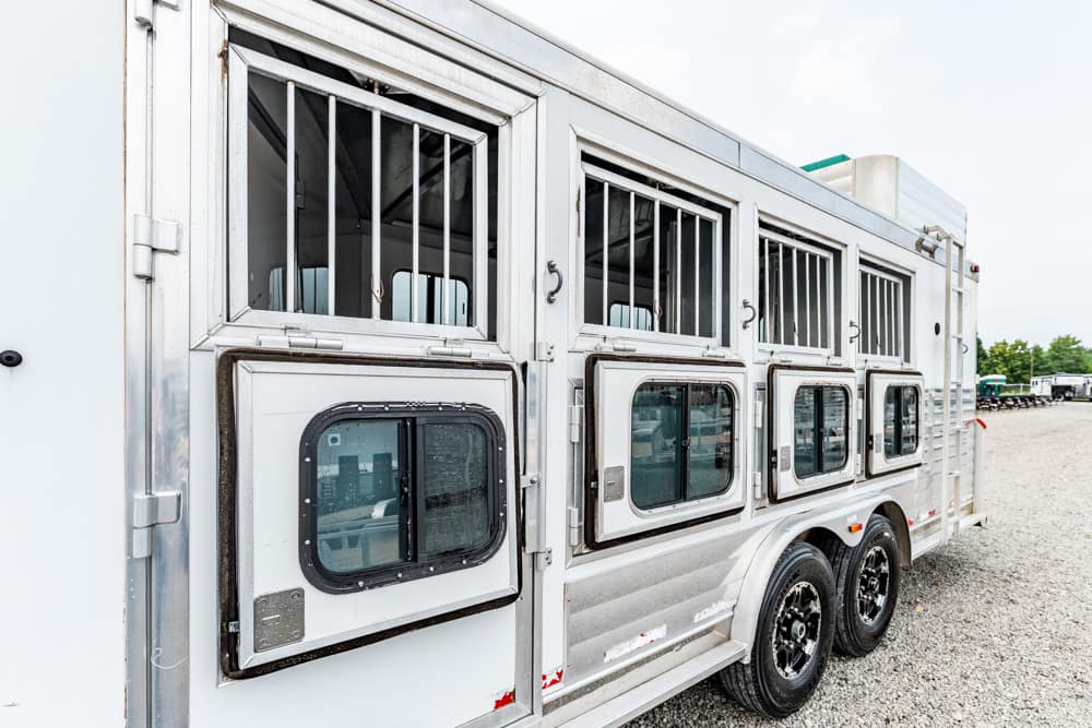 Silver horse trailer with multiple stalls and barred upper windows on a gravel lot.