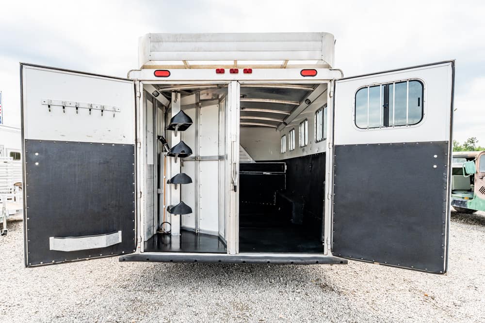 Rear of a white and black horse trailer with doors wide open, showing empty stalls and tack racks on the left wall.