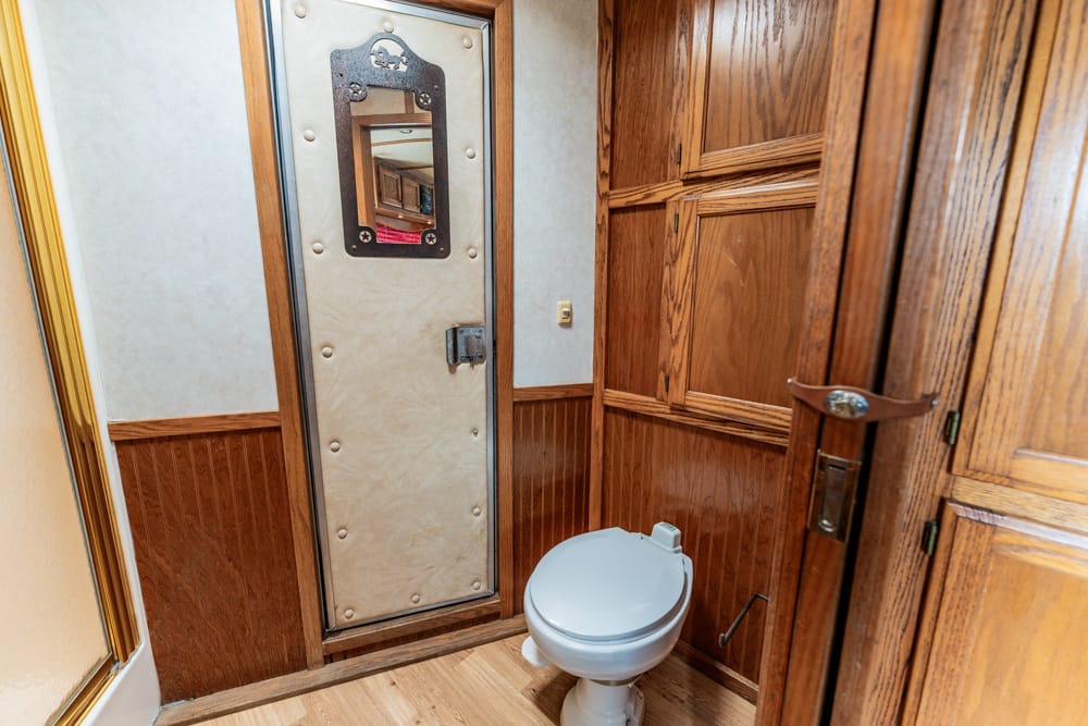 Narrow bathroom with wood paneling, a gray toilet, and a tall beige metal door with a window and heavy bolts.