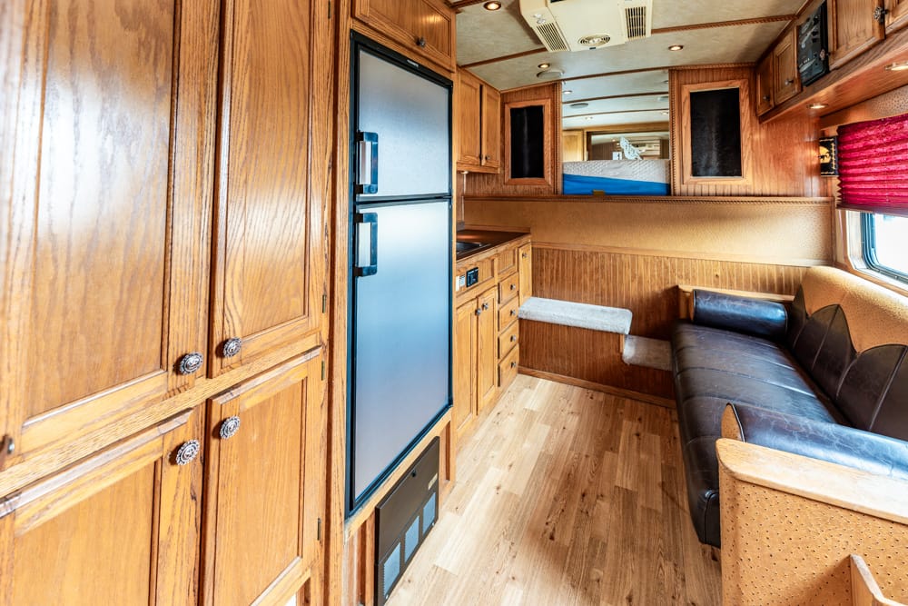 Wood-paneled RV interior with built-in fridge, abundant cabinets, and a black leather sofa along a windowed wall.