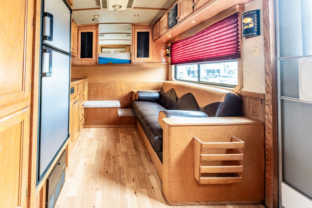Wood-paneled RV interior with a brown leather sofa, kitchen cabinetry, and a red window shade.