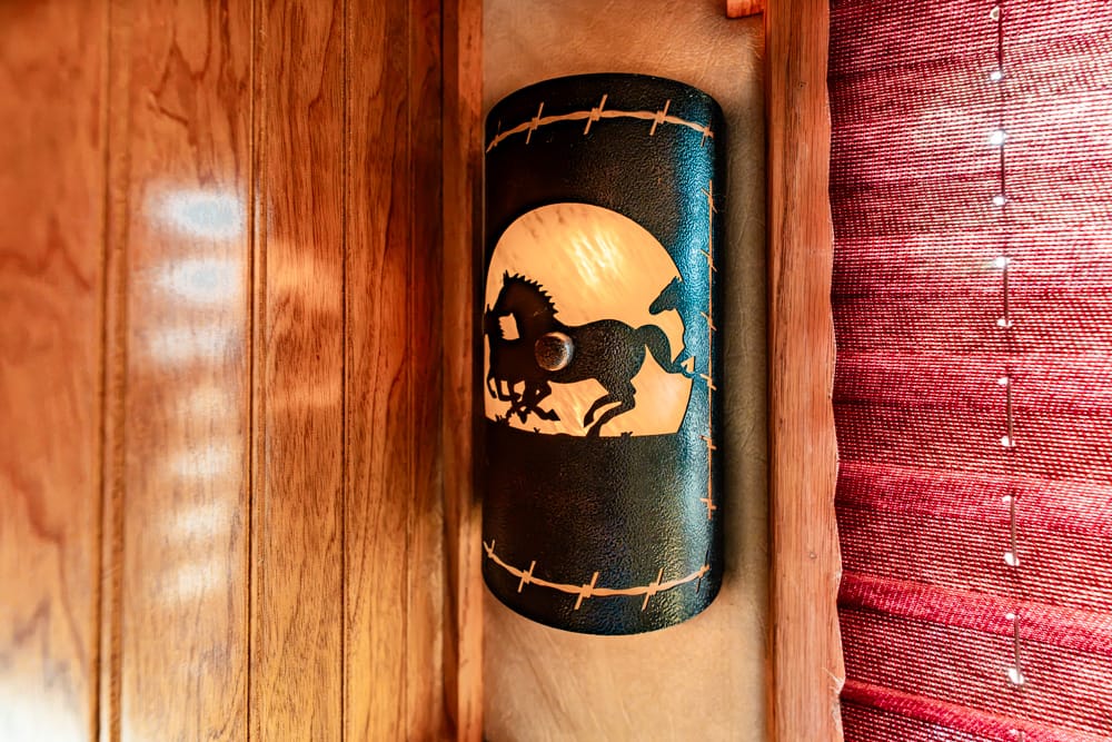 Decorative black cylindrical wall plaque with a bucking horse and rider silhouette in a yellow circle, framed by barbed-wire design, mounted on wood paneling.