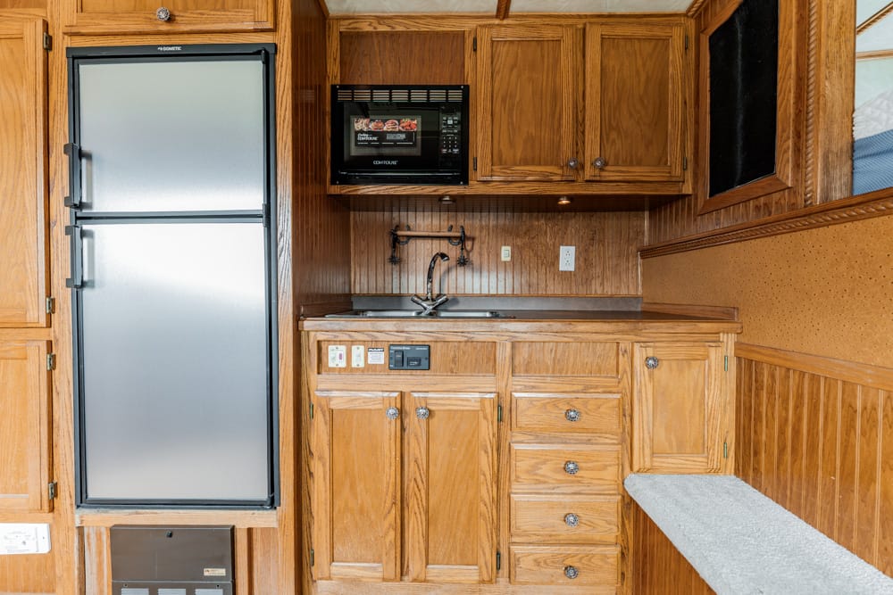 Compact wooden kitchen area with a stainless refrigerator on the left, microwave above the counter, sink, and multiple cabinets and drawers.