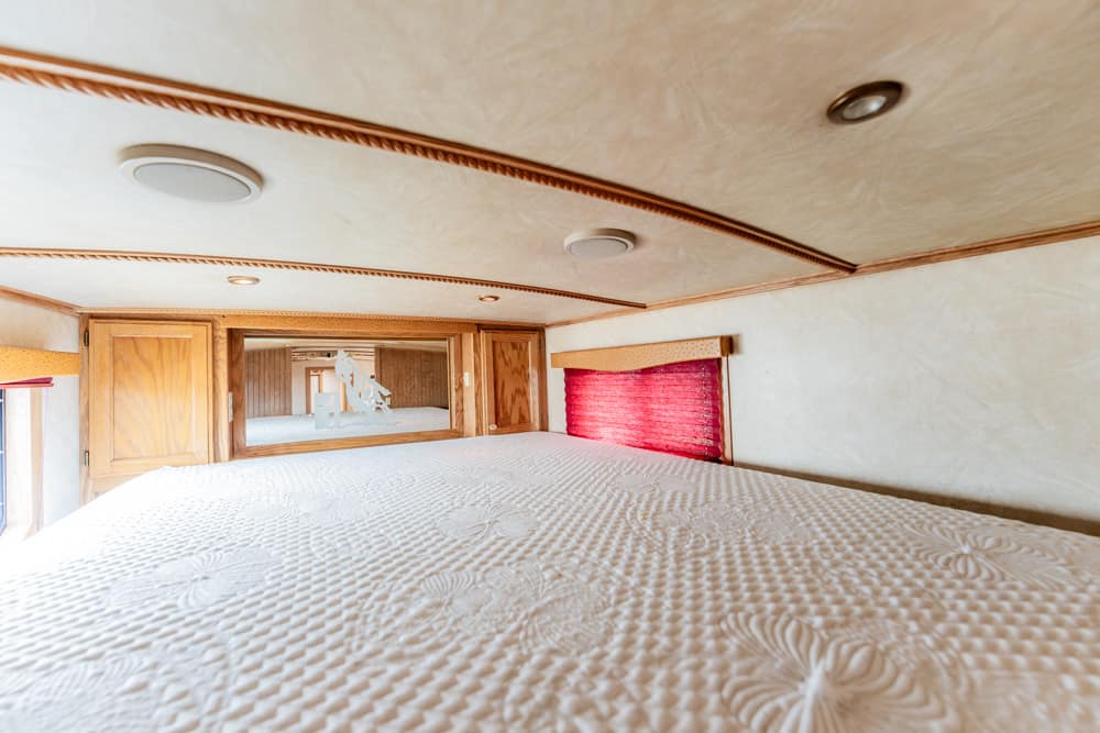 Cozy RV bedroom with a large bed, wooden cabinets above, and a mirrored wall reflecting more of the small space; a red window shade on the right adds color.