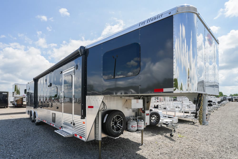 Glossy black toy hauler fifth-wheel trailer parked on a gravel lot, with spare tire and propane tanks beneath the front section against a blue sky.