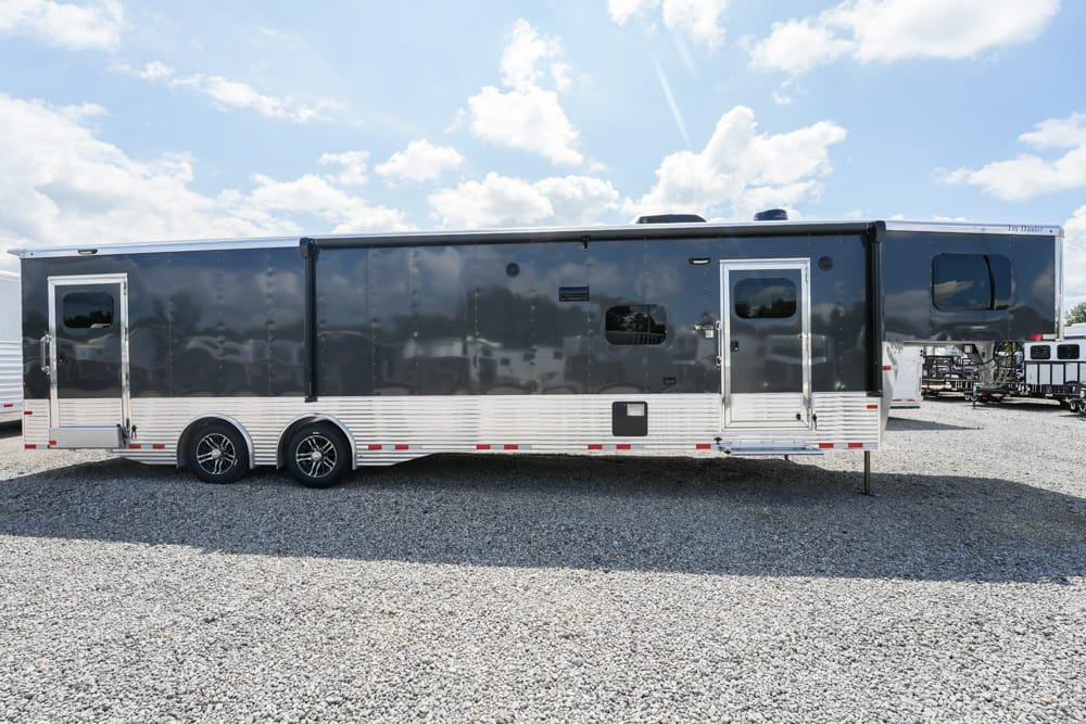 Long dark blue travel trailer with silver lower trim and two axles parked on gravel under a blue sky with scattered clouds.