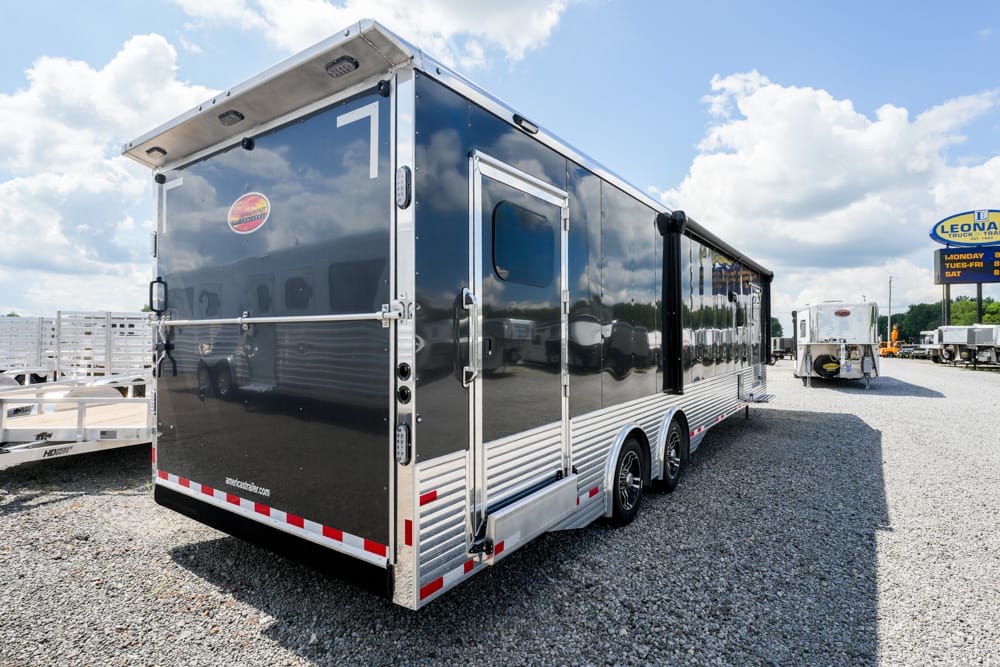 Long black multi-axle trailer with a glossy aluminum lower trim parked in a gravel lot, reflecting its surroundings in the sunlight.